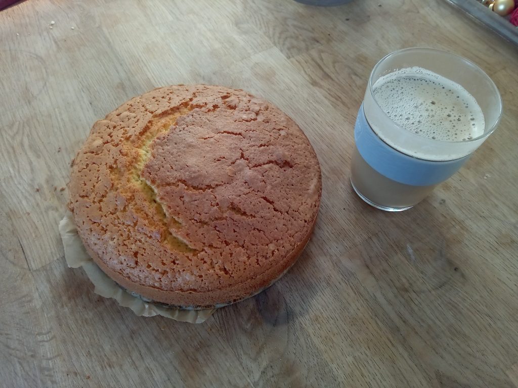 Ein kleiner Eierlikörkuchen steht auf einem Holztisch, daneben ein Glas Milchkaffee