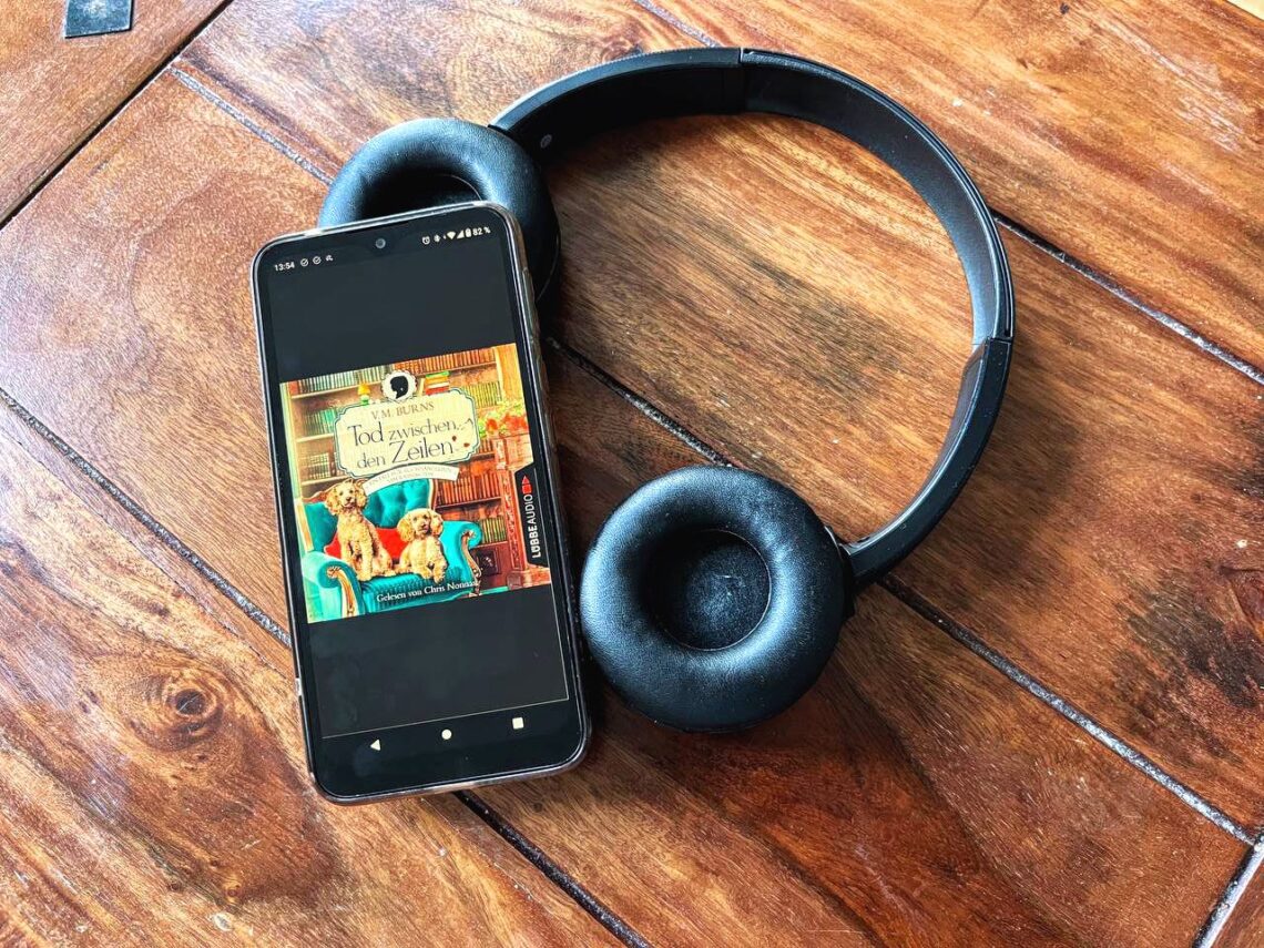 "Tod zwischen den Zeilen" von V. M. Burns auf einem Handy, daneben ein paar Bluetooth-Kopfhörer
